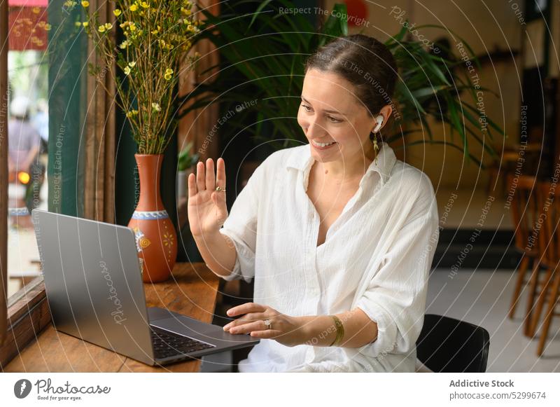 Lächelnde Frau winkt mit der Hand während eines Videogesprächs am Laptop Videoanruf Video-Chat Wellenhand abgelegen Arbeit Gruß freiberuflich Café Vietnam
