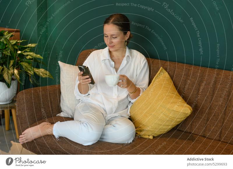 Lächelnde Frau, die in einem Café auf ihrem Smartphone surft und Kaffee trinkt Cappuccino Kaffeepause benutzend trinken Tee heiter Pause heiß Getränk Mobile