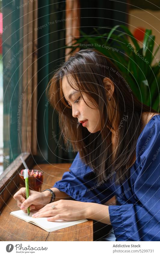 Konzentrierte asiatische Frau schreibt in einem Café in einen Notizblock zur Kenntnis nehmen Notebook Schüler lernen trinken Konzentration schreiben Bildung