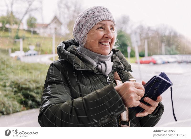Fröhliche ältere Frau mit Brieftasche auf der Straße Lächeln positiv Glück Großstadt heiter urban Hut reif warme Kleidung Freude Senior Outfit Lifestyle Stadt