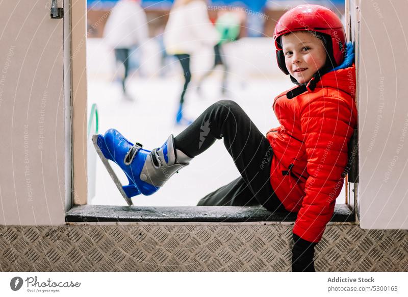Junge in Schlittschuhen sitzt am Rande einer Eisbahn Winter Sicherheit Schritt Kind lernen Freizeit Sport Aktivität Hobby kalt Schnee Fokus Erholung aktiv