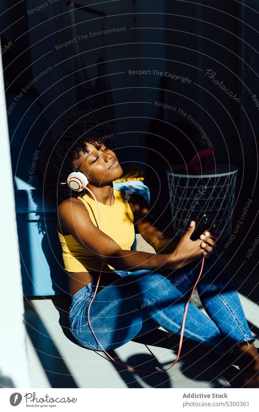 Zufriedene schwarze Frau mit Kopfhörern und Smartphone, die im Sonnenlicht chillt Musik zuhören heimwärts sich[Akk] entspannen Kälte genießen benutzend