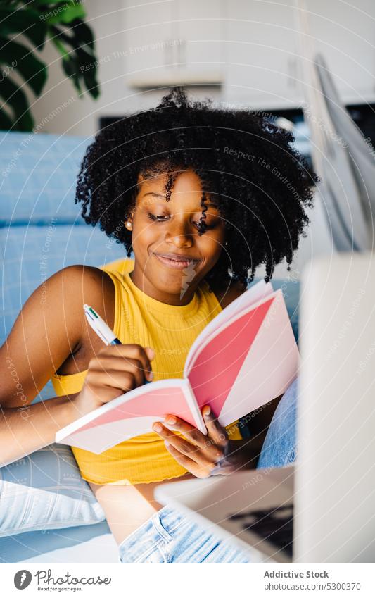 Zufriedene schwarze Frau macht sich Notizen in einem Notizblock zur Kenntnis nehmen Lächeln heiter schreiben heimwärts Notebook Planer Tagebuch Hinweis Sofa