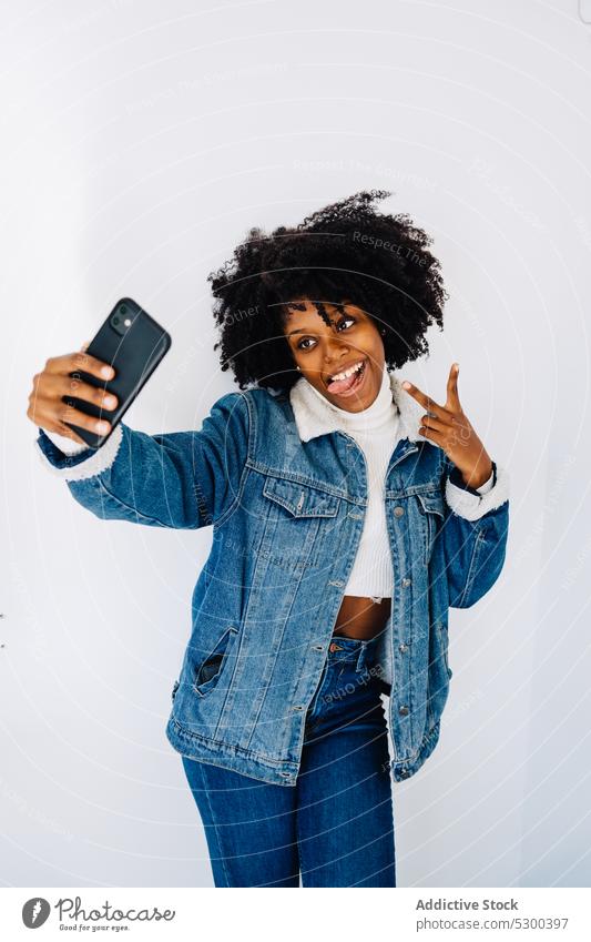 Fröhliche schwarze Frau macht Selfie Smartphone Selbstportrait benutzend Zunge zeigen Afro-Look Grimasse heiter v-Zeichen Glück Afroamerikaner ethnisch Frisur
