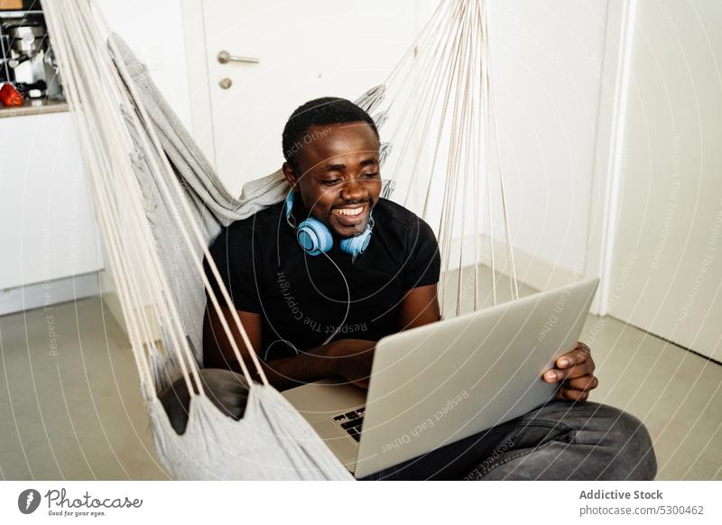 Positiver schwarzer Mann mit Kopfhörern am Laptop freiberuflich Lächeln Telearbeit benutzend Browsen heimwärts abgelegen männlich Afroamerikaner heiter ethnisch
