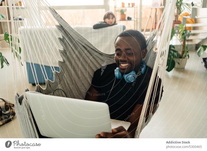 Positiver schwarzer Mann mit Kopfhörern am Laptop freiberuflich Lächeln Telearbeit benutzend Browsen heimwärts abgelegen männlich Afroamerikaner heiter ethnisch