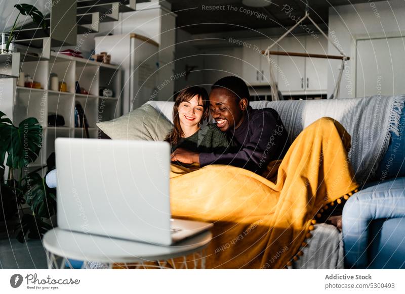 Vielfältiges Paar, das einen Film auf dem Laptop ansieht zuschauen kuscheln Zusammensein Partnerschaft Decke Umarmung heimwärts rassenübergreifend multiethnisch