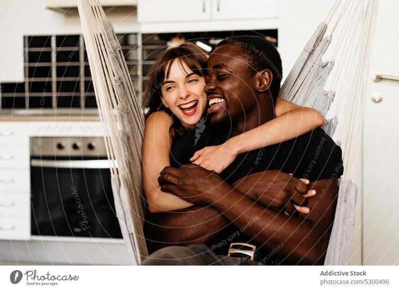 Glückliches multiethnisches Paar, das sich auf einem Schaukelstuhl umarmt Lachen Zusammensein kuscheln Küche Umarmung Wochenende heiter Kälte