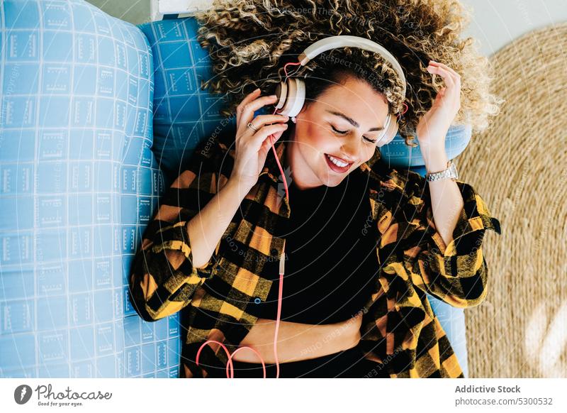 Fröhliche Frau mit Kopfhörern auf dem Sofa liegend zuhören Musik zu Hause Lügen Melodie Klang Lächeln positiv jung krause Haare Inspiration Information