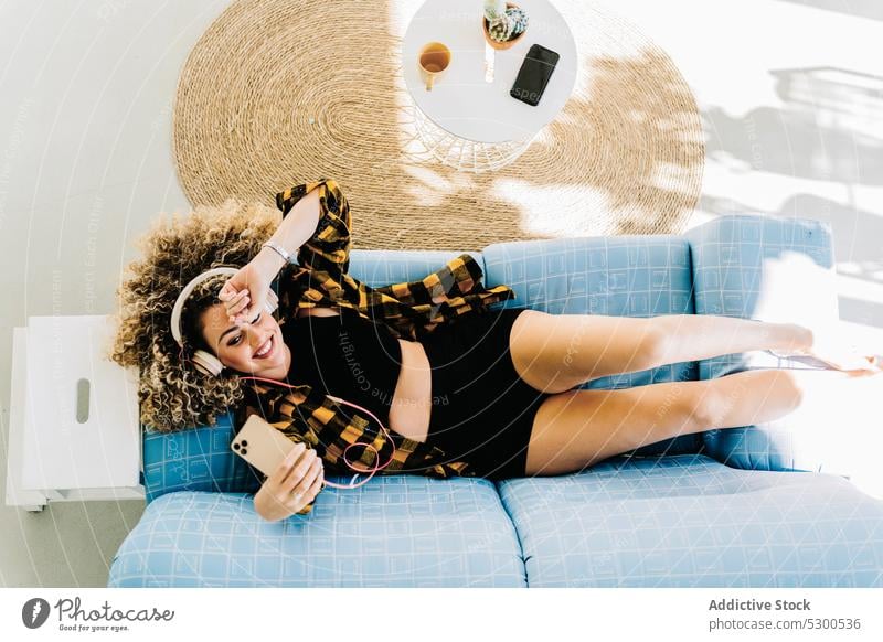 Fröhliche Frau mit Kopfhörern, die ein Selfie mit ihrem Smartphone macht benutzend zuhören Musik zu Hause Lügen Sofa Lächeln positiv jung krause Haare Drahtlos