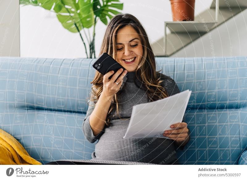 Lächelnde schwangere Frau mit Dokumenten im Telefongespräch benutzend Smartphone Telefonanruf Schriftstück Appartement reden Gespräch Sofa jung Arbeit