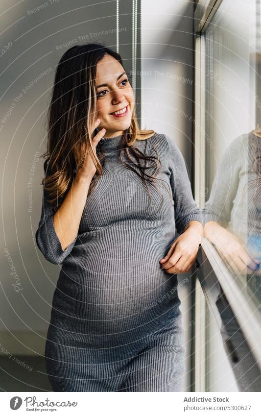 Lächelnde schwangere Frau, die mit einem Handy telefoniert benutzend Smartphone sprechen Fenster Telefonanruf Appartement Gespräch Kommunizieren positiv jung