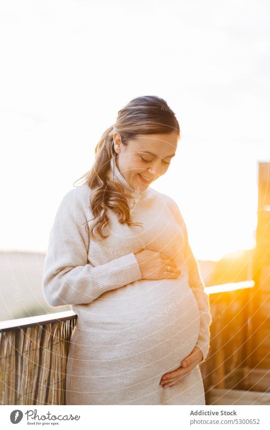 Glückliche schwangere Frau, die ihren Bauch auf der Terrasse umarmt ungeboren Baby Angebot Liebe Streicheln Bauch anfassen Sonnenlicht Lächeln Sonnenuntergang
