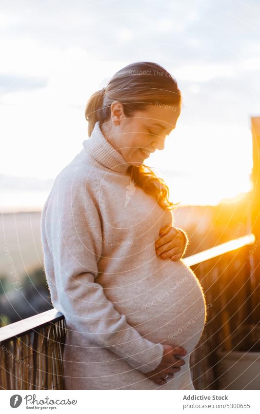 Glückliche schwangere Frau, die ihren Bauch auf der Terrasse umarmt ungeboren Baby Angebot Liebe Streicheln Bauch anfassen Sonnenlicht Lächeln jung