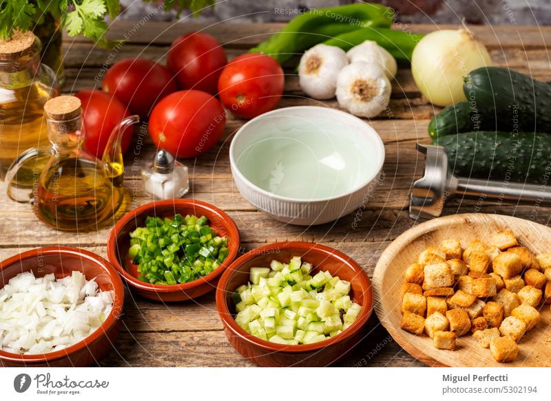 Die notwendigen Zutaten für eine Gazpacho um eine leere Schüssel herum, und mit dem gehackten Gemüse und dem gerösteten Brot zum Übergießen auf einem rustikalen Holztisch.