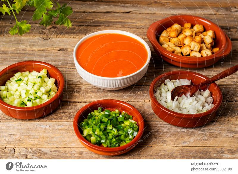 Schale mit Gazpacho neben einigen Tontellern mit gehacktem Gemüse und geröstetem Brot zum Gazpacho, auf einem rustikalen Tisch. Schalen & Schüsseln Zutaten