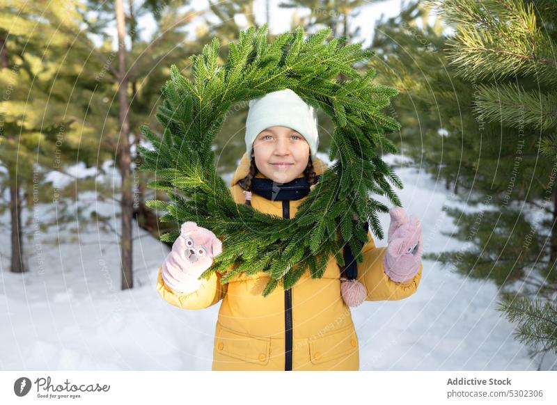 Niedliches Mädchen mit Kranz im verschneiten Wald Winter Natur Schnee Totenkranz warme Kleidung Lächeln nadelhaltig Hut kalt Wälder Saison Oberbekleidung Glück