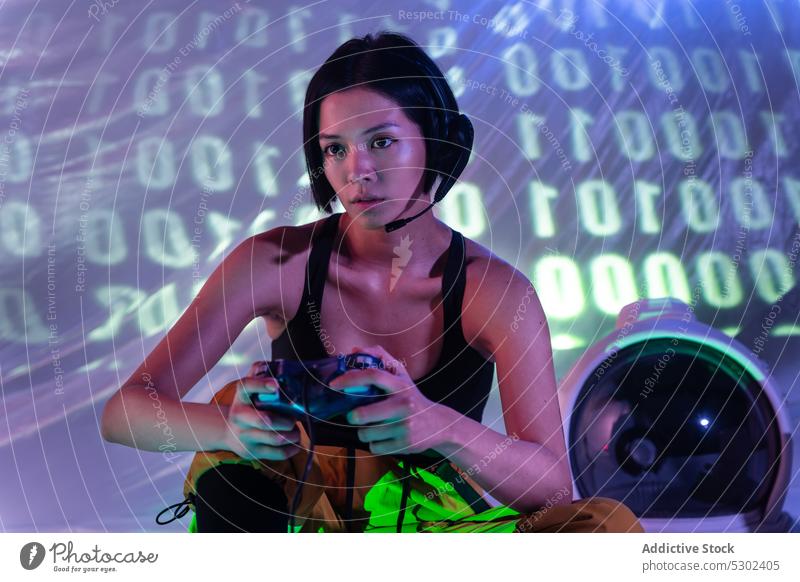 Selbstbewusste Frau mit Joystick im Neonlicht spielen Videospiel Spieler Schutzhelm Kopfhörer neonfarbig Apparatur unterhalten Gerät sitzen ernst jung Konsole