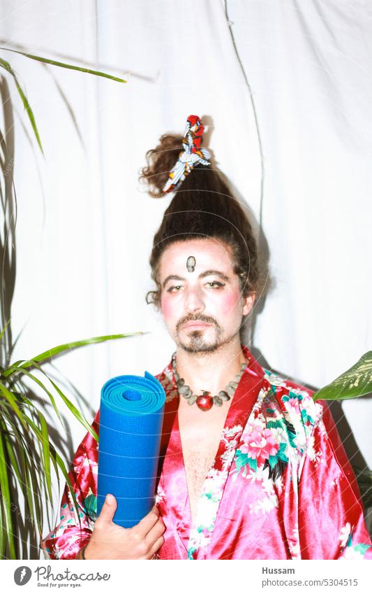 ist ein Fotokonzept über einen Mann, der eine Yogamatte hält, ein geblümtes Kleid trägt und seine Haare auf eine sehr lustige Weise gestylt hat. Sein Make-up sieht aus wie eine Geisha mit einem ironischen Blick.