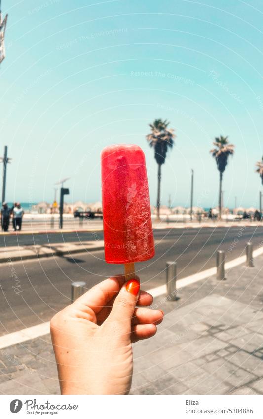 Hand hält rotes Eis am Stiel an einer Strandpromenade im Urlaub. Sommer verrotten lecker strandpromenade Erfrischung sommerlich Palmen kalt gefroren süß