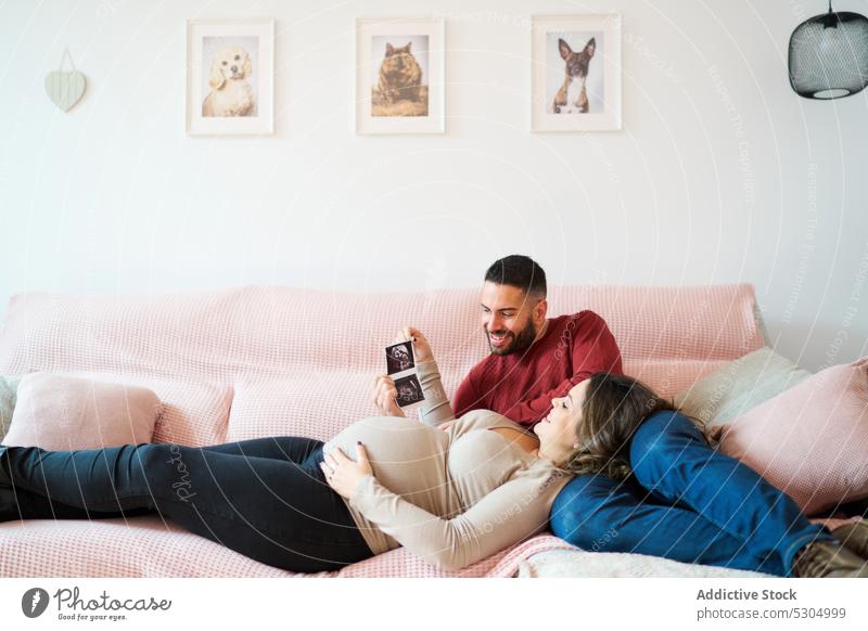 Lächelndes schwangeres Paar betrachtet Ultraschall zu Hause Schwangerschaft zuschauen Liebe Baby Sofa Zusammensein Ehefrau Ehemann Glück Partnerschaft Liege