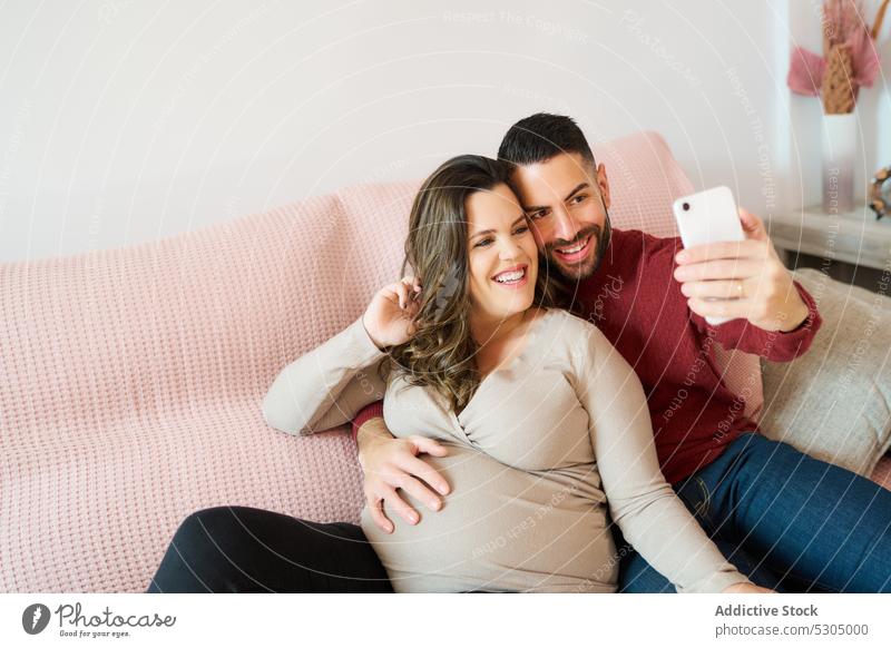 Glückliches schwangeres Paar nimmt Selfie auf Handy Smartphone Zusammensein Partnerschaft Liebe benutzend Bonden Schwangerschaft heiter Sofa Wohnzimmer Lächeln