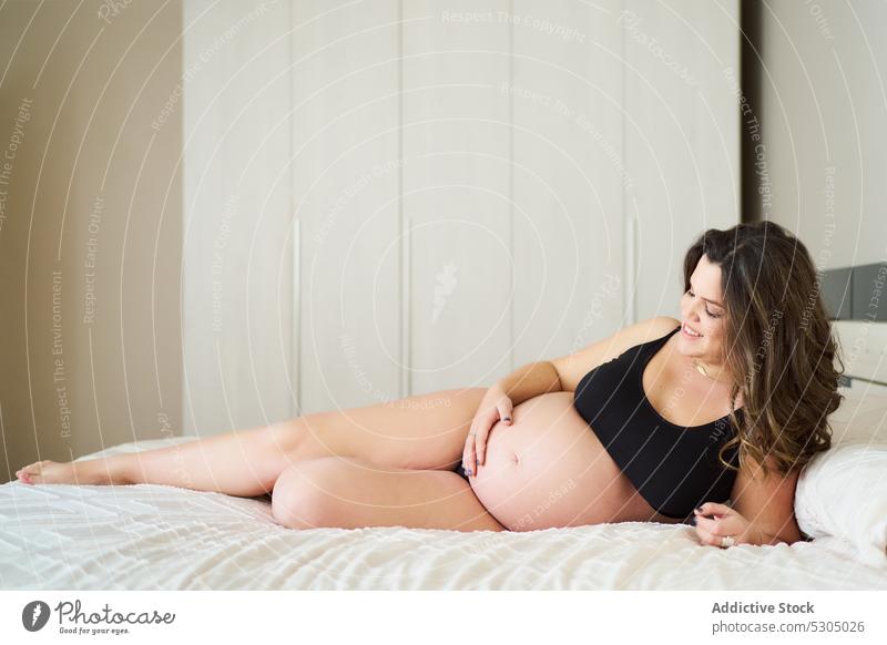 Lächelnde schwangere Frau auf dem Bett liegend Glück Bauch anfassen mütterlich Schlafzimmer Unterwäsche erwarten Lügen positiv jung Kopfkissen Komfort berühren