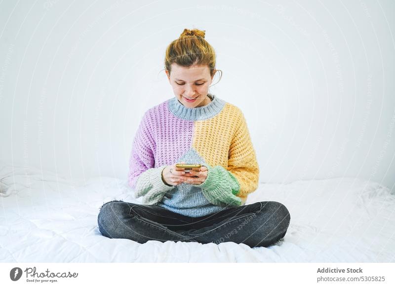 Fröhliche Frau, die auf dem Bett auf ihrem Smartphone surft benutzend heimwärts Lächeln positiv Browsen Glück Gerät Mobile Apparatur Nachricht Pullover jung