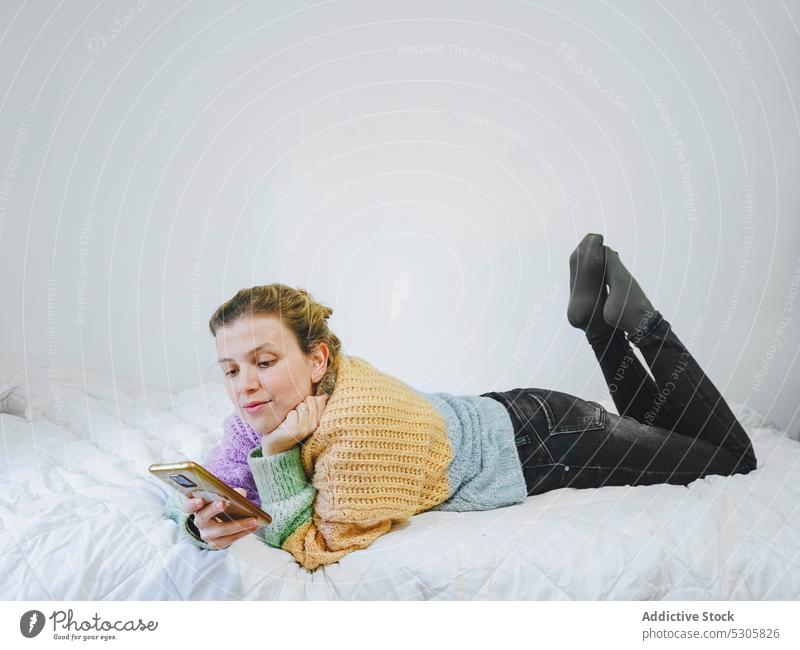 Fröhliche Frau, die auf dem Bett auf ihrem Smartphone surft benutzend heimwärts Lügen Lächeln positiv Browsen Glück Gerät Mobile Apparatur Nachricht Pullover