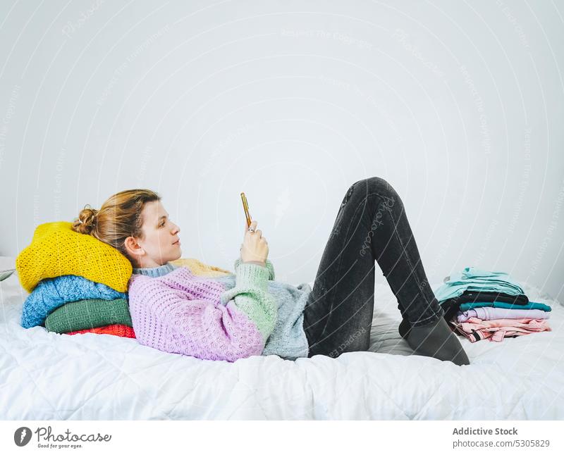 Frau surft auf dem Bett auf ihrem Smartphone benutzend heimwärts Lügen Lächeln positiv Browsen Glück Gerät Mobile Apparatur Nachricht Pullover jung padmasana