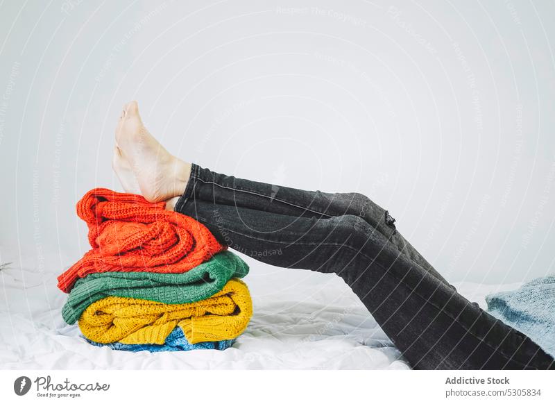 Anonyme Person mit Beinen auf einem Stapel warmer Pullover farbenfroh Jeanshose Lügen Stoff Bett Garn Haufen Strickwaren heimwärts Gewebe weich Textil Komfort