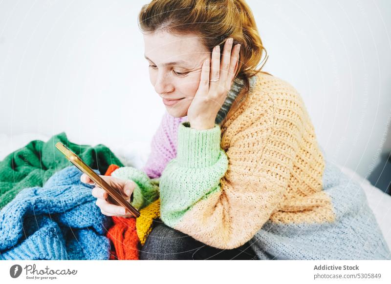 Zufriedene Frau surft auf Smartphone in der Nähe eines Kleiderstapels Lächeln Bett Nachricht benutzend Browsen soziale Netzwerke Glück positiv Pullover Mobile