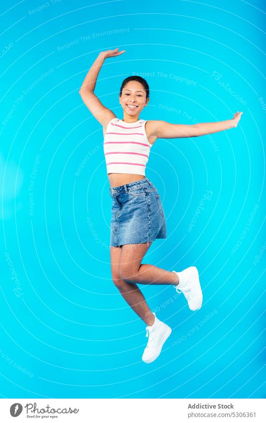 Fröhliche junge ethnische Dame springt in blauem Studio Frau springen Lächeln Arme hochgezogen aufgeregt Energie heiter Model Freude positiv Outfit Glück Stil