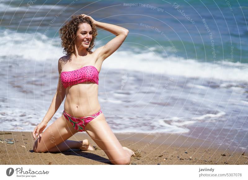 Glückliche Frau im Bikini auf den Knien sitzend am Sandstrand MEER Strand Haare berühren Lächeln knien Sommer Urlaub krause Haare Badebekleidung Küste Resort