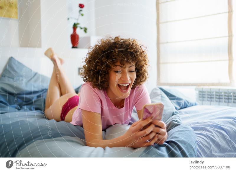 Aufgeregte Frau, die ein Handy benutzt, während sie zu Hause im Bett liegt benutzend Smartphone erstaunt aufgeregt zuschauen Video Lügen Browsen heimwärts