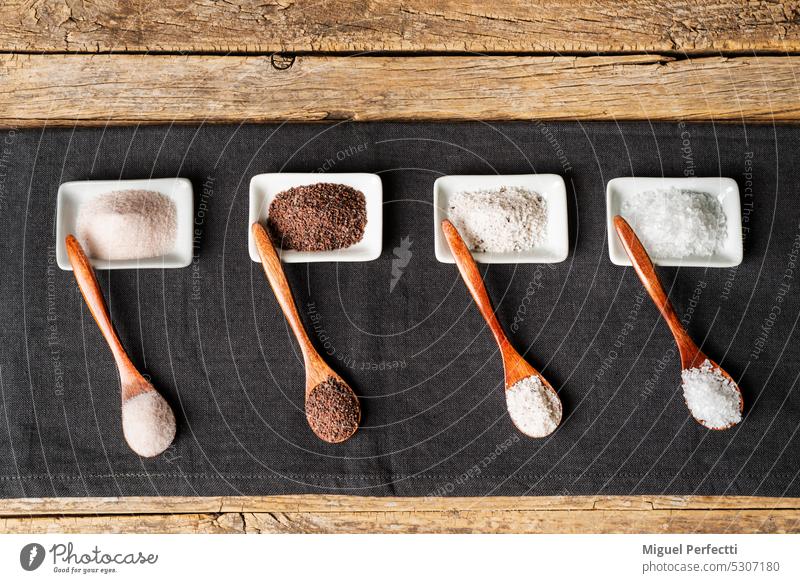 Holzlöffel neben einigen kleinen Tellern mit verschiedenen Arten von Salz, schwarz, rosa, Flocken und gewürzt mit Trüffel, auf einem Tischtuch auf einem rustikalen hölzernen Hintergrund.top view.