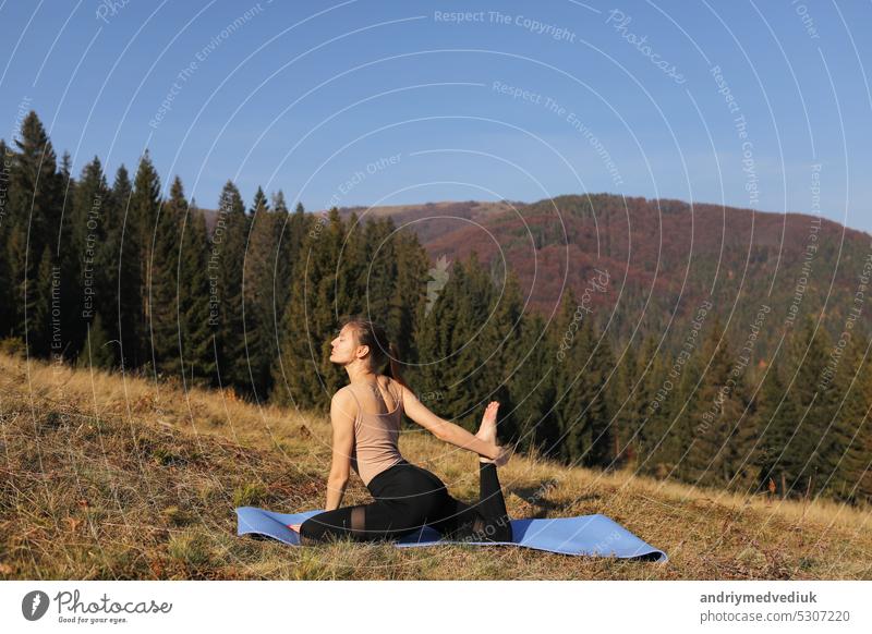 Junge fitte Frau macht Übungen auf Yoga-Matte auf dem Hintergrund der sonnigen Berge Hügel. Outdoor-Workout, gesunder Lebensstil. Frau im Sportanzug dehnt sich in der Natur gegen den Wald am Morgen