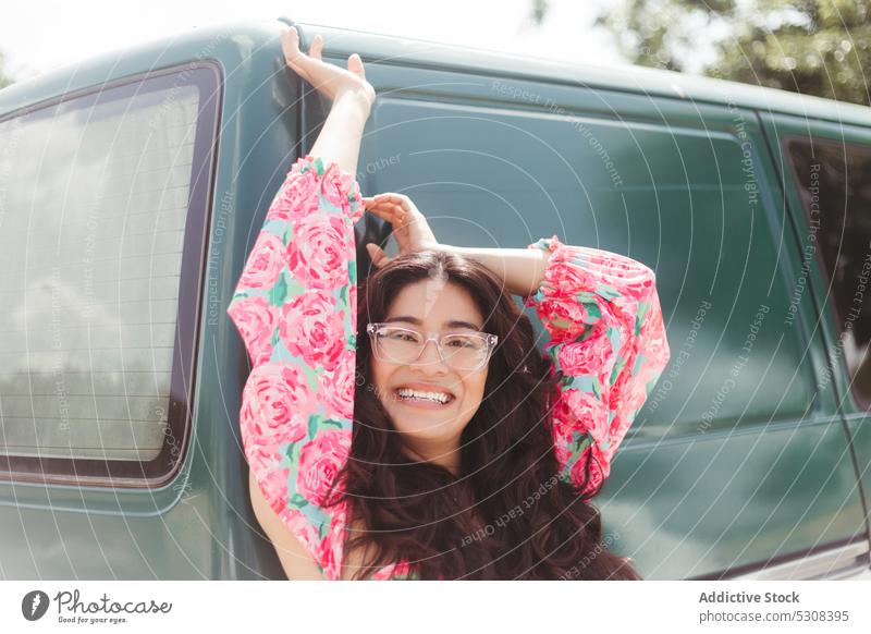 Lächelnde Frau steht vor einem olivgrünen Auto PKW Sonnenlicht Sommer froh Brille heiter Optimist Inhalt sorgenfrei Glück Freude jung Freiheit genießen Fahrzeug