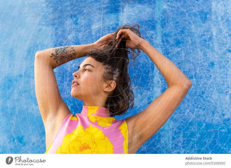 Verträumte Frau berührt Haar gegen gemalte blaue Wand nachdenklich Straße Windstille Haare berühren besinnlich farbenfroh Stil trendy Großstadt Tattoo urban