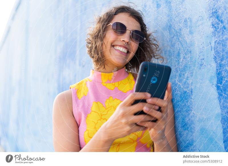 Fröhliche Frau, die auf der Straße ihr Smartphone durchsucht benutzend Nachricht Lächeln farbenfroh Glück Sommer Stil heiter Sonnenbrille krause Haare jung