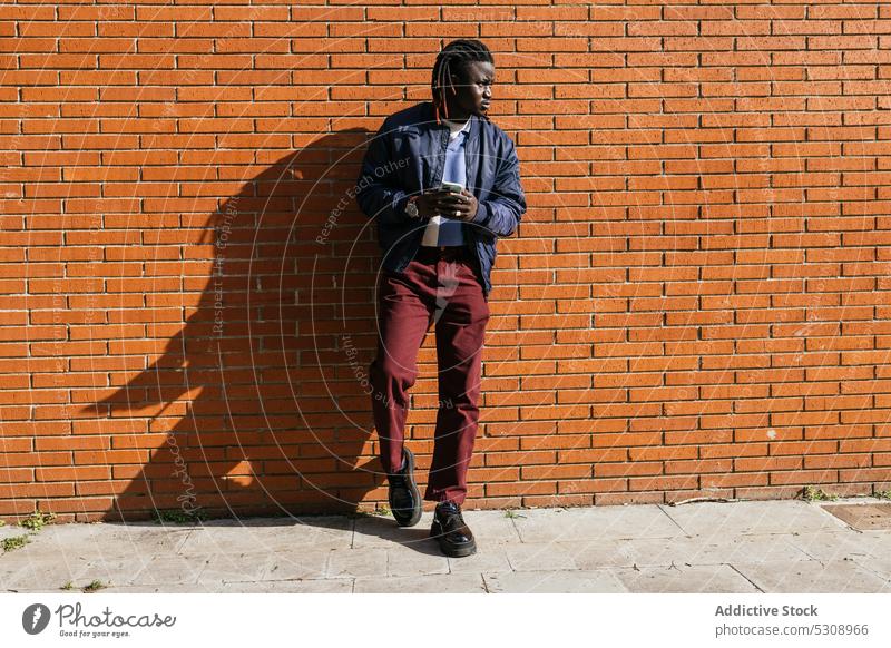 Schwarzer Mann mit Dreadlocks in der Nähe einer Backsteinmauer besinnlich nachdenklich Smartphone Mobile ernst Anschluss Browsen zuschauend Geflecht Rastalocken
