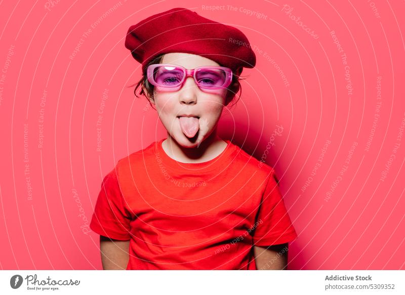 Lustiges Kind in rotem Freizeitoutfit mit Brille und Baskenmütze Stil Mund geöffnet Zunge zeigen Gesicht machen Humor lustig Mädchen Model lebhaft farbenfroh