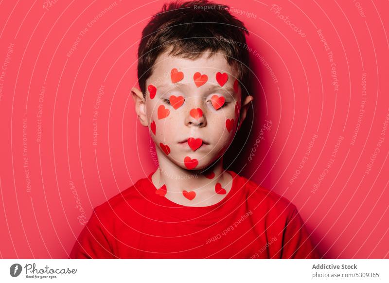 Niedlicher Junge in rotem Freizeithemd mit rotem Liebesaufkleber im Gesicht beim Posieren für ein Foto Kind Atelier niedlich Aufkleber lässig modern bezaubernd