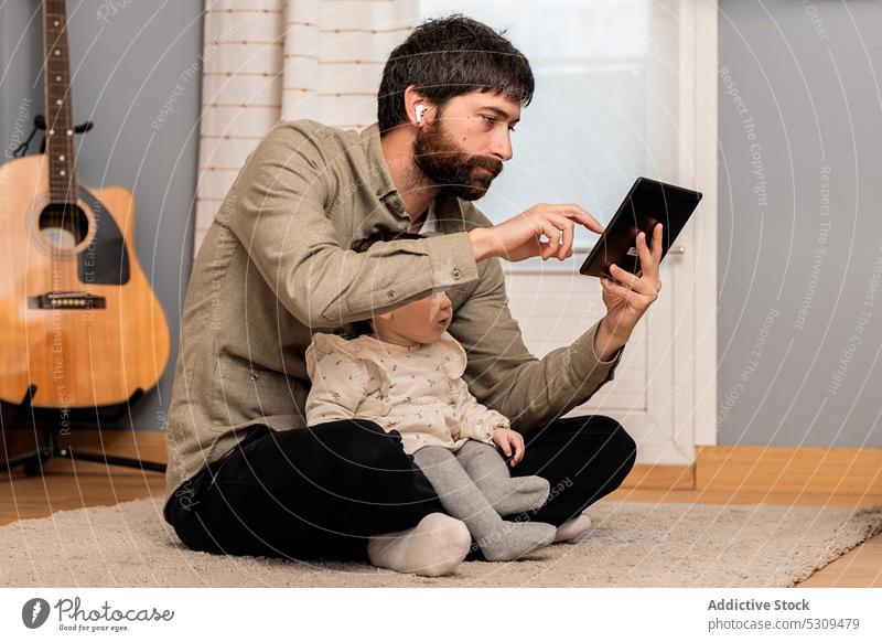 Neugieriges Mädchen schaut auf den Bildschirm eines Tablets neben einem Mann, der auf dem Boden sitzt Tablette Vater Tochter benutzend abgelegen heimwärts