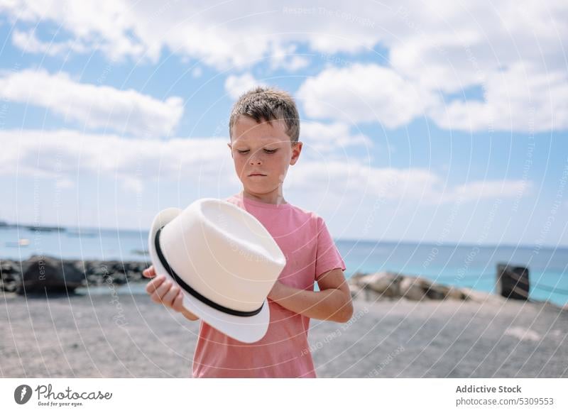 Seriöser Junge mit Hut steht bei Tageslicht am Meer Blauer Himmel MEER Sommer selbstbewusst Kind Haltehut Natur ernst jung Kindheit Lifestyle Strand sorgenfrei
