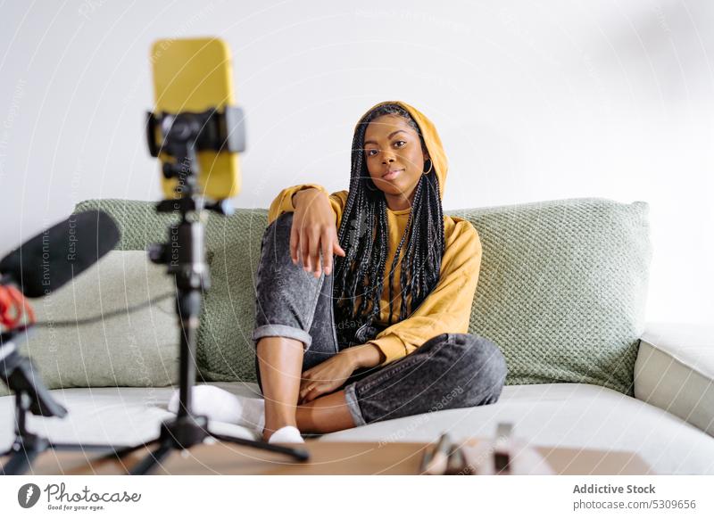Zufriedene schwarze Frau im Kapuzenpulli, die auf dem Sofa ein Video aufnimmt Blogger Smartphone Aufzeichnen Podcast Browsen benutzend Laptop vlog