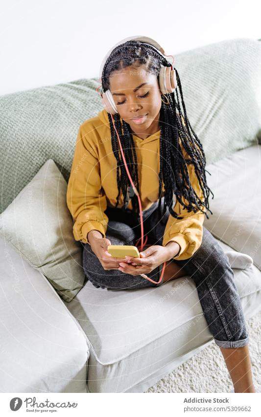 Ruhige schwarze Frau, die auf dem Sofa auf ihrem Smartphone surft und Musik hört zuhören benutzend heimwärts Kopfhörer Browsen Surfen Afroamerikaner Apparatur