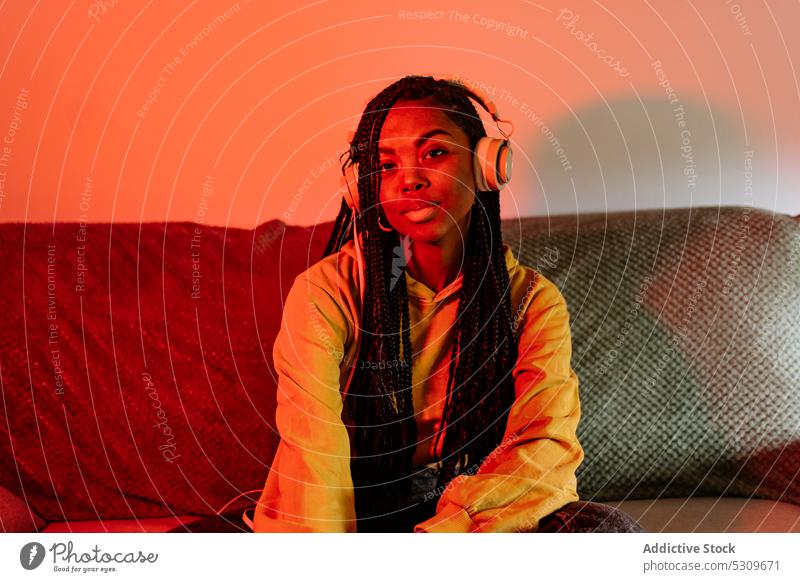 Charmante afroamerikanische Frau, die auf dem Sofa mit Kopfhörern Musik hört zuhören neonfarbig ernst heimwärts ruhen Gesang meloman Geflecht Apparatur jung