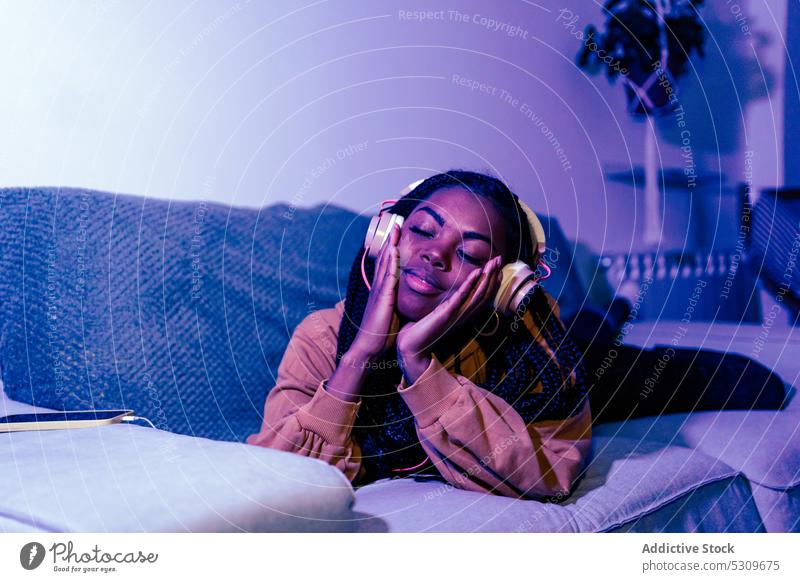 Entspannte schwarze Frau, die mit geschlossenen Augen auf dem Sofa Musik hört zuhören Kopfhörer neonfarbig Lügen heimwärts sich[Akk] entspannen Windstille