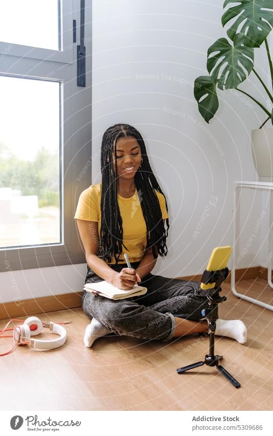 Fokussierte schwarze Frau schreibt Notizen in ein Copybook, während sie sich auf die Aufnahme eines Videos vorbereitet Smartphone Blogger Lächeln Aufzeichnen